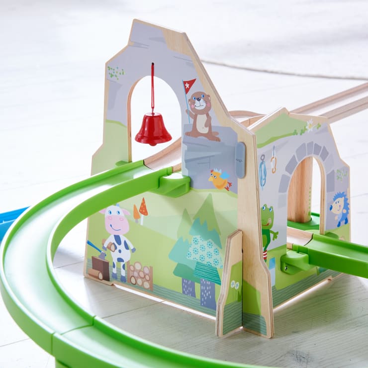 Ein Spielzeugeisenbahntunnel aus Holz mit illustrierten Tieren, darunter eine Kuh, ein Frosch, ein Otter und ein Igel. Grüne Kunststoffschienen schlängeln sich durch den Tunnel, und in einem der Bögen hängt eine rote Glocke.