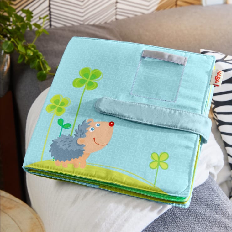 Ein weiches Kinderbuch aus Stoff mit einem Cartoon-Igel, grünen Kleeblättern und einem blauen Hintergrund liegt auf einer Couch. Das Buch hat ein Stoffband und ist mit Klettverschluss befestigt.