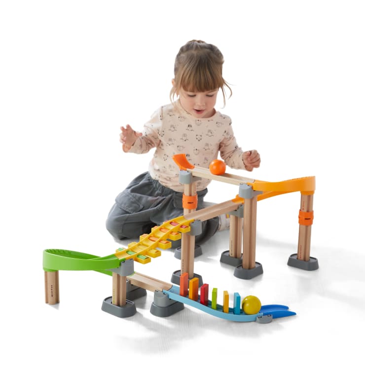 Ein kleines Kind sitzt auf dem Boden und spielt mit einer bunten Holzmurmelbahn mit Schienen, Stufen und Rampen. Zwei Murmeln sind auf den Bahnen zu sehen. Der Hintergrund ist weiß.