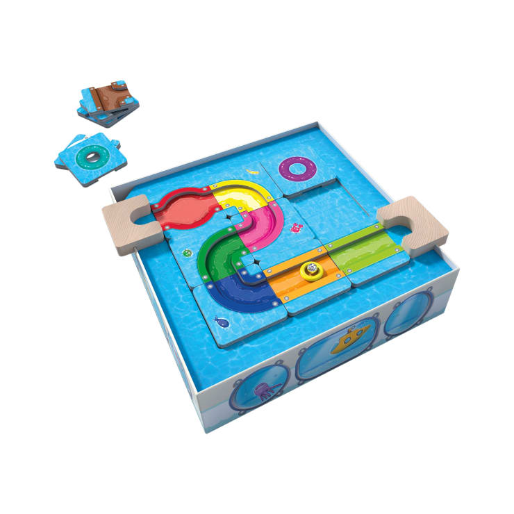Ein Murmelbahnspiel für Kinder mit bunten gebogenen und geraden Bahnen in einer blauen Box, die an Wasser erinnert. Zwei zusätzliche Puzzleteile befinden sich außerhalb der Schachtel auf weißem Hintergrund.