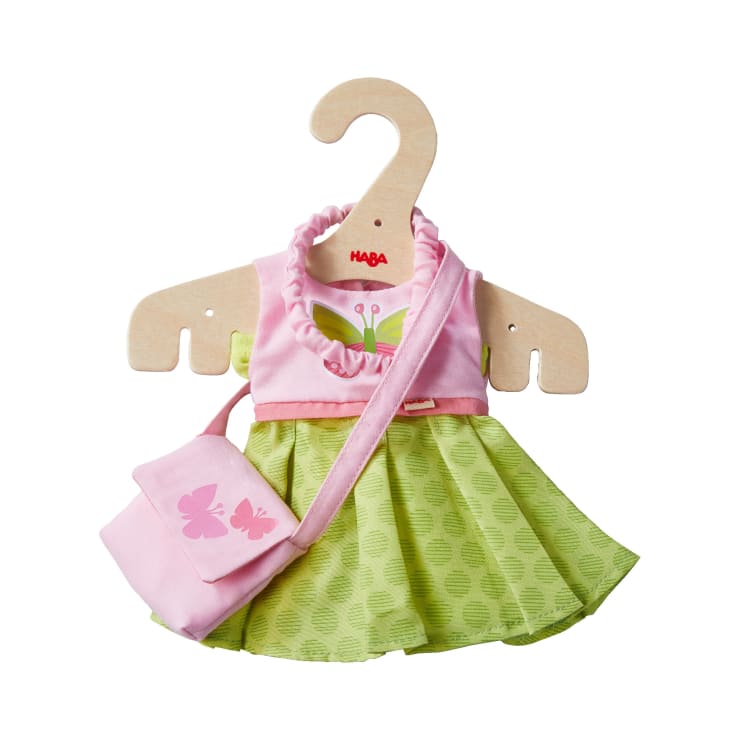 An einem Holzbügel hängt ein Puppenkostüm, bestehend aus einem rosa-grünen Kleid mit Schmetterlingsmuster und einer passenden rosa Umhängetasche, ebenfalls mit Schmetterlingen verziert.