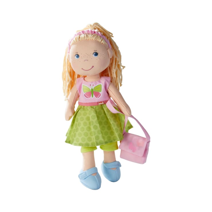 Eine weiche Stoffpuppe mit blondem Garnhaar, die ein rosafarbenes Oberteil mit einem Schmetterling, einen grünen Rock, grüne Leggings, blaue Schuhe und ein rosafarbenes Stirnband trägt und eine rosafarbene Handtasche in der Hand hält, auf weißem Hintergrund.