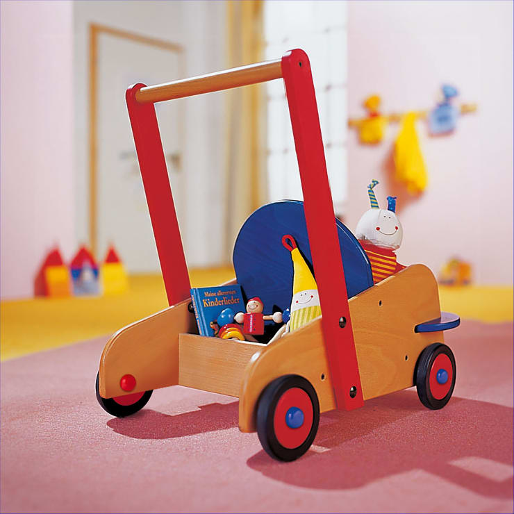 Ein hölzerner Spielzeugwagen mit roten Griffen und Rädern, blauen Akzenten und einer Ablagefläche für kleine Spielsachen und ein Kinderbuch auf einem rosa Teppich in einem hell erleuchteten Raum.
