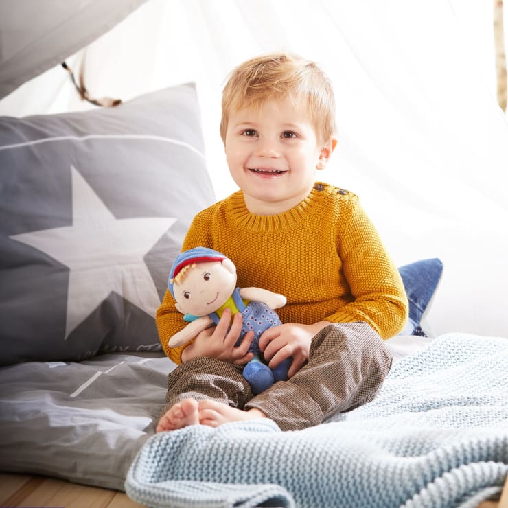 Ein kleines Kind mit kurzen blonden Haaren, das einen senfgelben Pullover und eine karierte Hose trägt, sitzt auf einem Bett mit grauer und blauer Bettwäsche, hält eine weiche Puppe und lächelt in die Kamera.