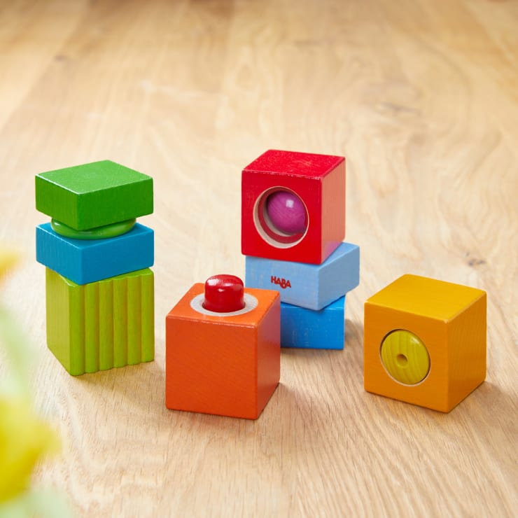 Vier bunte, stapelbare Holzklötze, jeder mit einer anderen Farbe und einem anderen Design, sind auf einer hellen Holzoberfläche angeordnet. Einige Blöcke haben runde Objekte im Inneren oder oben, die durch kreisförmige Ausschnitte sichtbar sind.