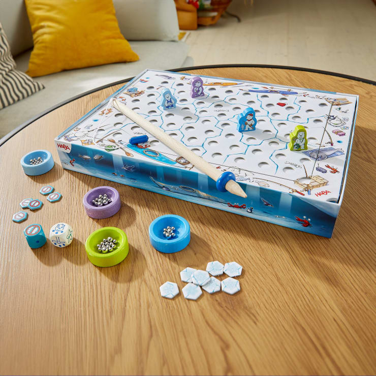 Ein Brettspiel mit einer Angelrute, farbigen Spielsteinen, Murmeln, Würfeln und sechseckigen Spielsteinen ist auf einem Holztisch in einem Wohnzimmer mit einer Couch, Kissen und einem gelben Kissen im Hintergrund aufgebaut.