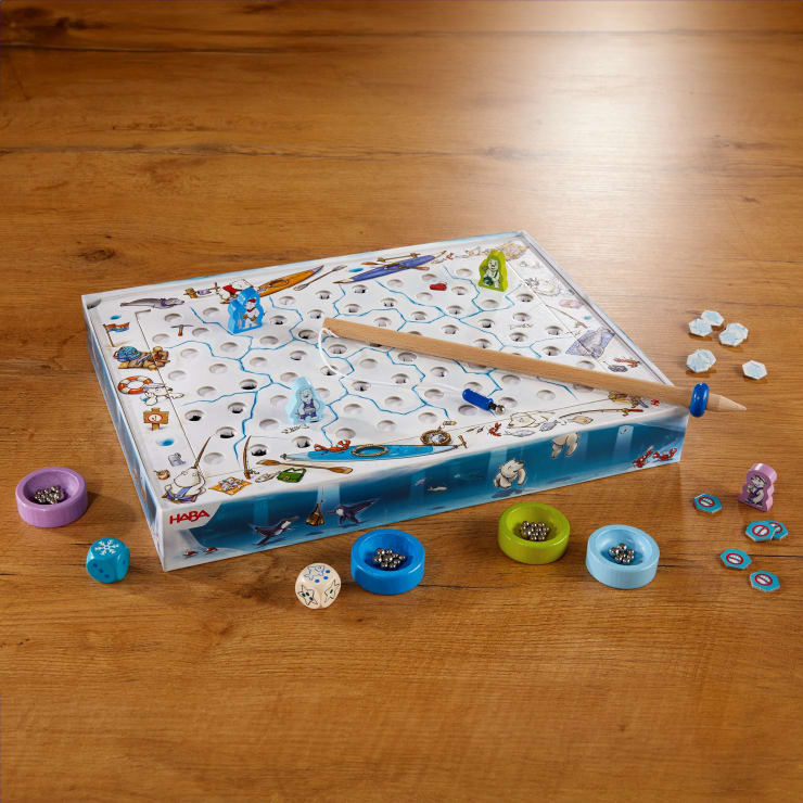 Ein Brettspiel mit einem winterlichen Thema wird auf einem Holztisch aufgebaut. Das Spiel besteht aus einer Schachtel mit vielen Löchern, farbigen Scheiben, Figuren, Würfeln, Spielsteinen und einem magnetischen Stab, der einer Angelrute ähnelt.