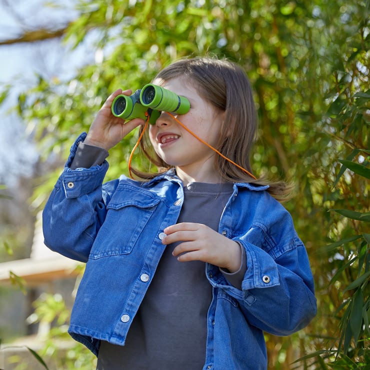 Ein Kind, das eine Jeansjacke trägt, schaut durch ein grünes Spielzeug-Fernglas im Freien, umgeben von grünem Laub und Sonnenlicht.