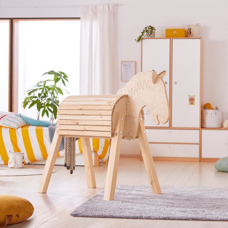 Ein hölzernes Steckenpferd steht in einem hellen, aufgeräumten Kinderzimmer mit weißen Wänden, einem Bett mit gelb gestreiftem Bettzeug, einer grünen Pflanze und einem weißen Kleiderschrank im Hintergrund.