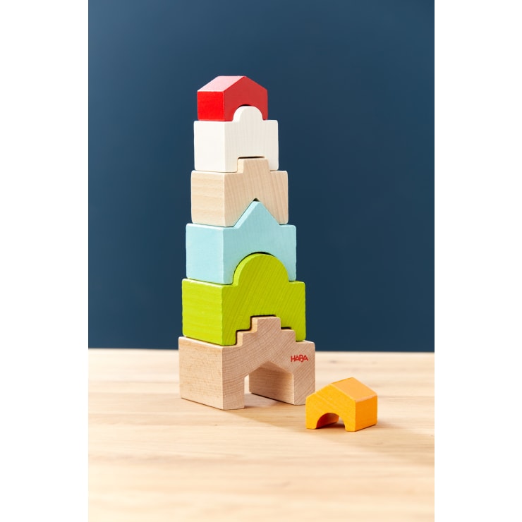 Ein Stapel von sieben bunten Holzklötzen in verschiedenen Formen und Größen liegt auf einer hellen Holzfläche mit dunkelblauem Hintergrund. Ein gelber Klotz befindet sich separat rechts vom Stapel.