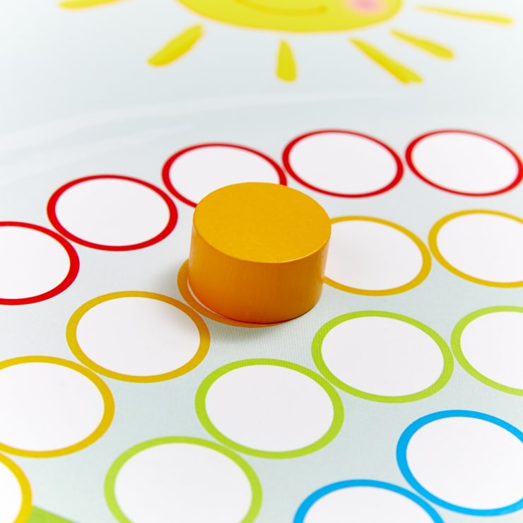 Ein gelber Zylinder steht auf einem Spielbrett mit leeren kreisförmigen Feldern, die in Rot, Gelb und Grün umrandet sind. Auf dem Hintergrund ist oben eine lächelnde Sonne abgebildet.