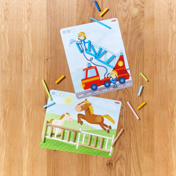 Zwei illustrierte Puzzletafeln auf einer hölzernen Oberfläche; eine zeigt Feuerwehrleute mit einem Feuerwehrauto, die andere ein Pferd, das über einen Zaun springt. Bunte Kreidestifte sind um die Puzzles herum verstreut.