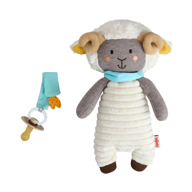 Ein Plüschschaf mit beiger und grauer Färbung, einem blauen Schal und hellbraunen Hörnern wird neben einem Babyschnuller mit blauem Clip und einem kleinen orangefarbenen Stoffteil gezeigt.