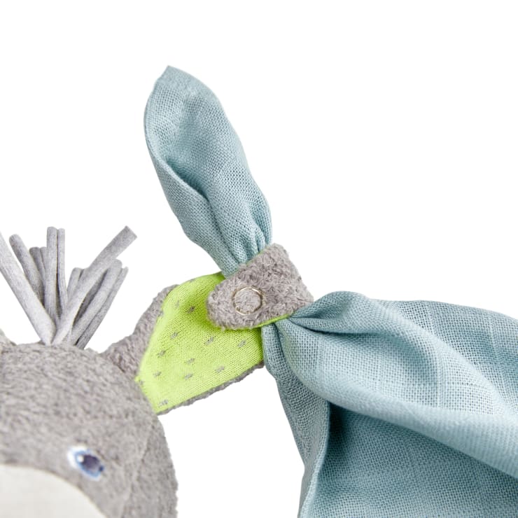 Eine Nahaufnahme eines Plüschtierohrs aus grauem und grünem Stoff, mit einem hellblauen Tuch darum gebunden und einem grauen Knopf, der den Knoten sichert. Das Plüschtier hat eine weiche, strukturierte Oberfläche.