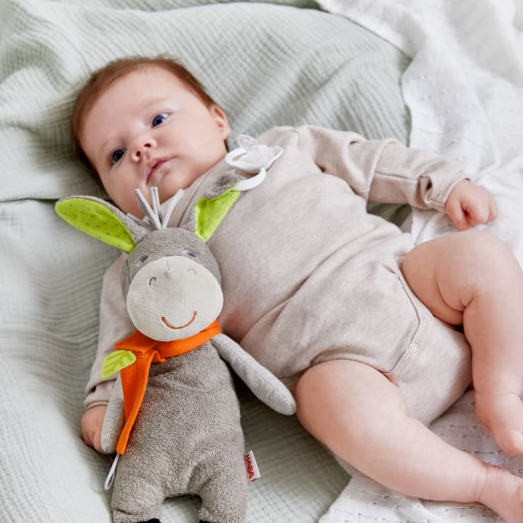 Ein Baby in einem beigen Strampler liegt auf einer weichen, hellen Decke neben einem grauen Plüschesel mit grünen Ohren und einem orangefarbenen Schal.