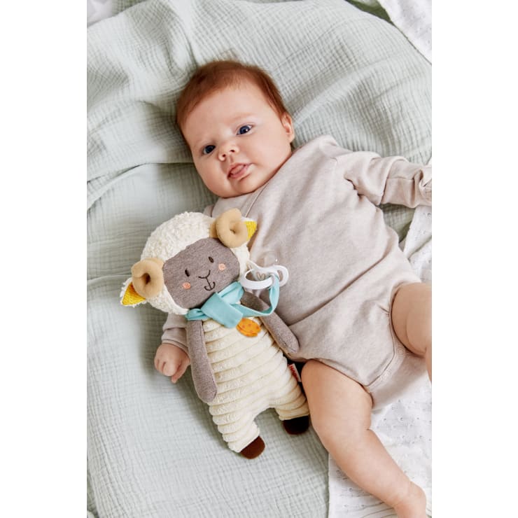 Ein Baby in einem beigen Strampler liegt auf einer hellgrünen Decke und hält ein Plüschschaf und einen Schnuller in der Hand. Das Baby blickt mit einem neutralen Gesichtsausdruck nach oben.