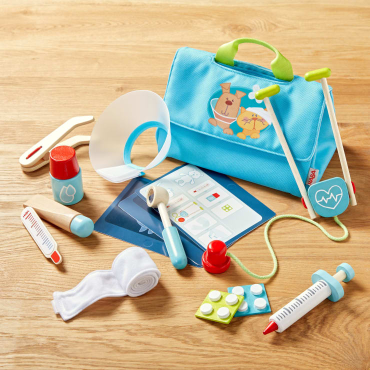 Eine blaue Spielzeug-Arzttasche mit einem Hunde- und Katzendesign ist von medizinischen Spielzeugen umgeben, darunter ein Stethoskop, ein Thermometer, ein Verband, Tablettenblister, eine Spritze, ein Reflexhammer und ein Klemmbrett auf einer Holzoberfläche.