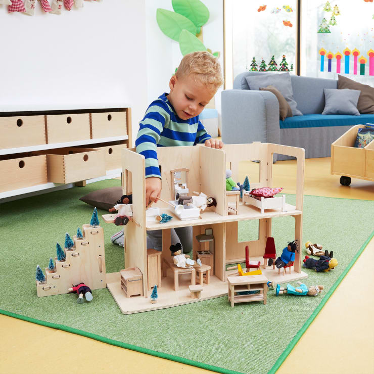 Ein kleines Kind in einem gestreiften Hemd spielt mit einem hölzernen Puppenhaus, das auf einem grünen Teppich steht. Im Inneren des Hauses sind kleine Puppen und Möbel aufgestellt. Der Raum hat natürliches Licht, Holzmöbel und Dekorationen an den Wänden.