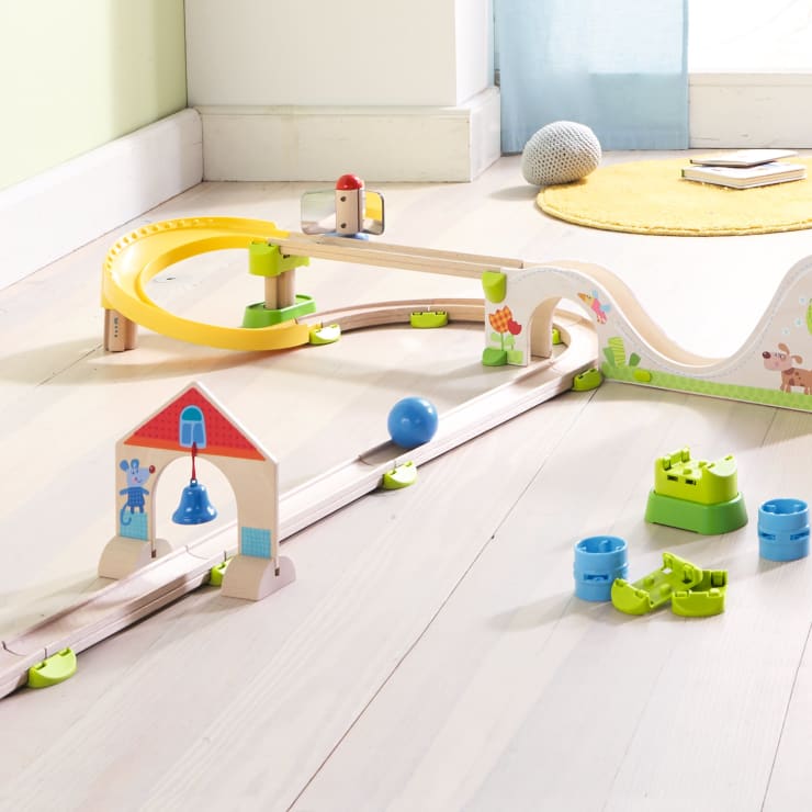 Ein hölzernes Murmelbahn-Spielzeugset ist auf einem hellen Holzboden mit Rampen, Kurven, einer Glocke, grünen Verbindungsstücken und einem blauen Ball aufgebaut. Ein gelber Teppich und ein graues Kissen sind in der Nähe eines Fensters im Hintergrund zu sehen.