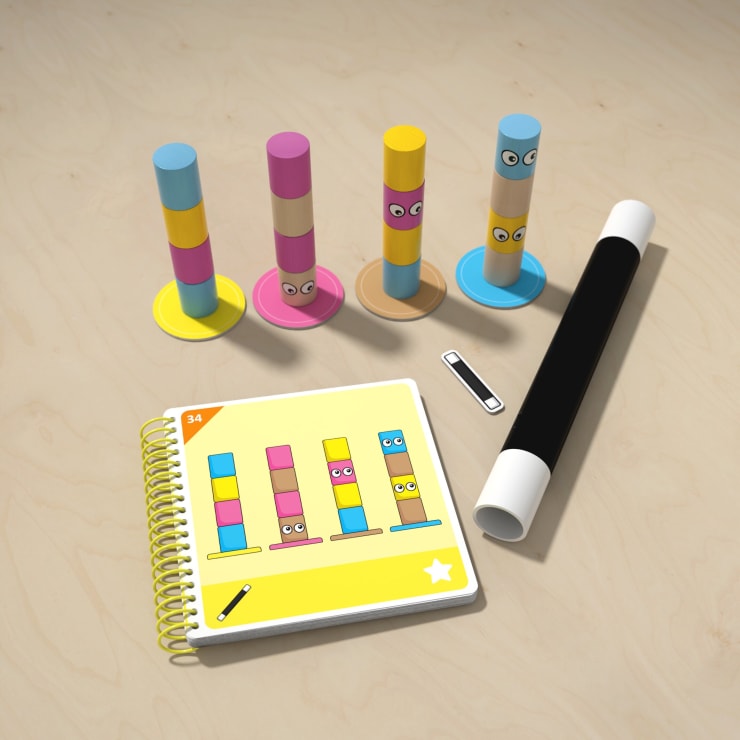 Vier bunte Stapelspielzeuge mit Gesichtern sind neben einem spiralgebundenen Activity Book angeordnet, das ein Stapelpuzzle zeigt. Ein schwarzer und weißer Marker und eine aufgerollte Matte befinden sich ebenfalls auf der hellen Holzoberfläche.