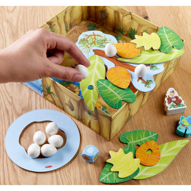 Eine Hand legt eine weiße eiförmige Figur auf ein buntes Brettspiel mit blattförmigen Figuren, einer Pappschachtel, weißen Eiern, einem blauen Ring, einem Würfel und tierischen Holzplättchen auf einer Holzoberfläche.