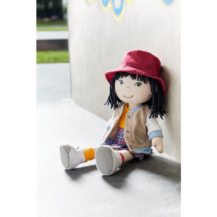 Eine Stoffpuppe mit schwarzem Garnhaar, die einen roten Hut, eine beige Weste, ein gelbes Hemd, einen karierten Rock und weiße Schuhe trägt, sitzt aufrecht an eine Betonwand gelehnt im Freien.