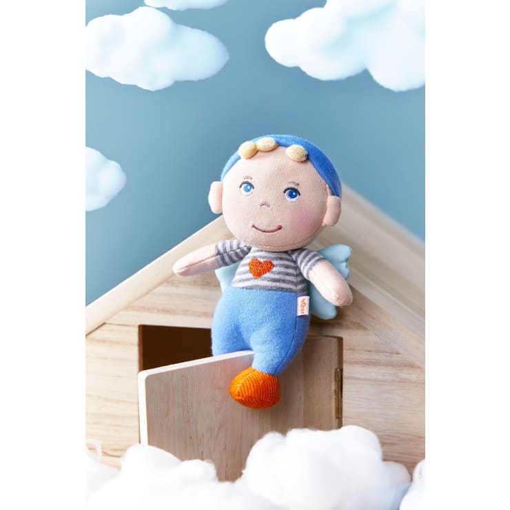 Eine Stofftierpuppe mit blauer Hose, gestreiftem Hemd und blauer Mütze sitzt am Rande eines Holzhauses. Im Hintergrund sind Wolken zu sehen, und baumwollartiger Stoff umgibt das Haus und schafft eine verträumte Atmosphäre.