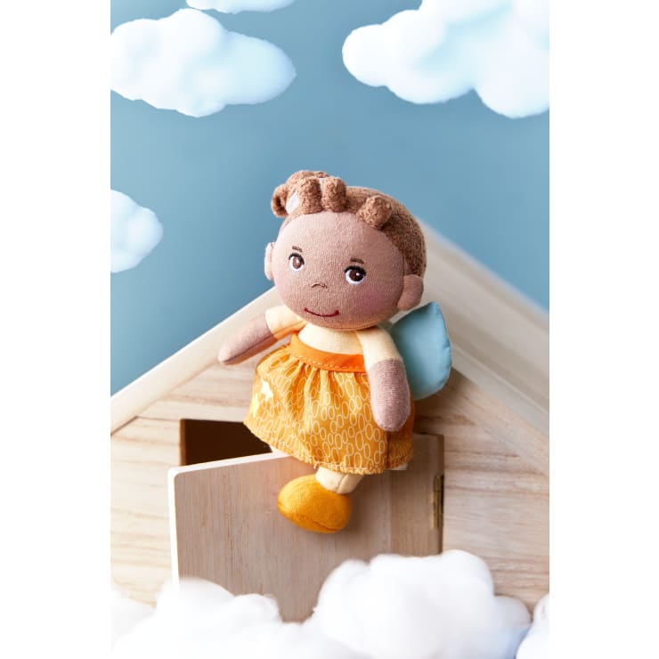 Eine Stoffpuppe mit braunem Haar, einem gelben Kleid und blauen Flügeln sitzt auf dem Dach eines kleinen Holzhauses, umgeben von flauschigen weißen Wolken und einem blauen Himmel im Hintergrund.