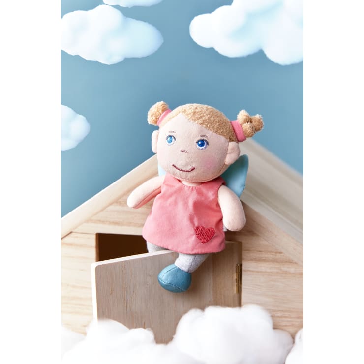 Eine Stoffpuppe mit blonden Zöpfen, blauen Augen, rosa Kleid und blauen Schuhen sitzt auf dem Rand eines Holzhauses mit offener Tür. Im Hintergrund sind flauschige Wolken und ein blauer Himmel zu sehen.