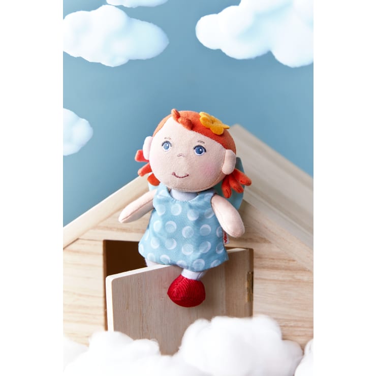 Eine Stoffpuppe mit roten Haaren, einem blauen gepunkteten Kleid und einer gelben Filzblume sitzt auf dem Dach eines kleinen Holzhauses, umgeben von Baumwollwolken und einem blauen Himmelshintergrund mit Wolkenausschnitten.