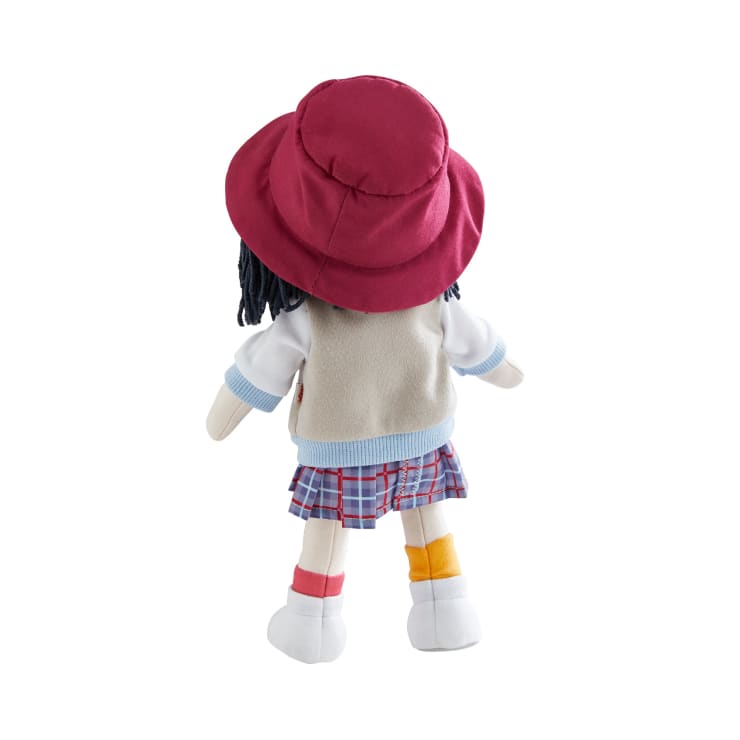 Eine Plüschpuppe, die mit dem Gesicht zur Wand steht und einen roten Hut, ein weißes Hemd mit beiger Weste, einen karierten Rock und verschiedenfarbige Socken - eine orangefarbene und eine rote - sowie weiße Schuhe trägt. Die Puppe hat dunkles Garnhaar und einen hellen Teint.