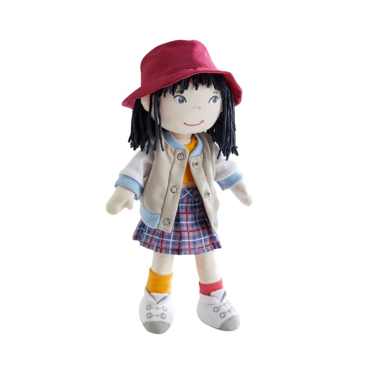 Eine Stoffpuppe mit schwarzem Garnhaar, die einen roten Hut, eine beige Jacke, ein gelbes Hemd, einen karierten Rock, verschiedene Socken und weiße Schuhe trägt und vor einem weißen Hintergrund steht.