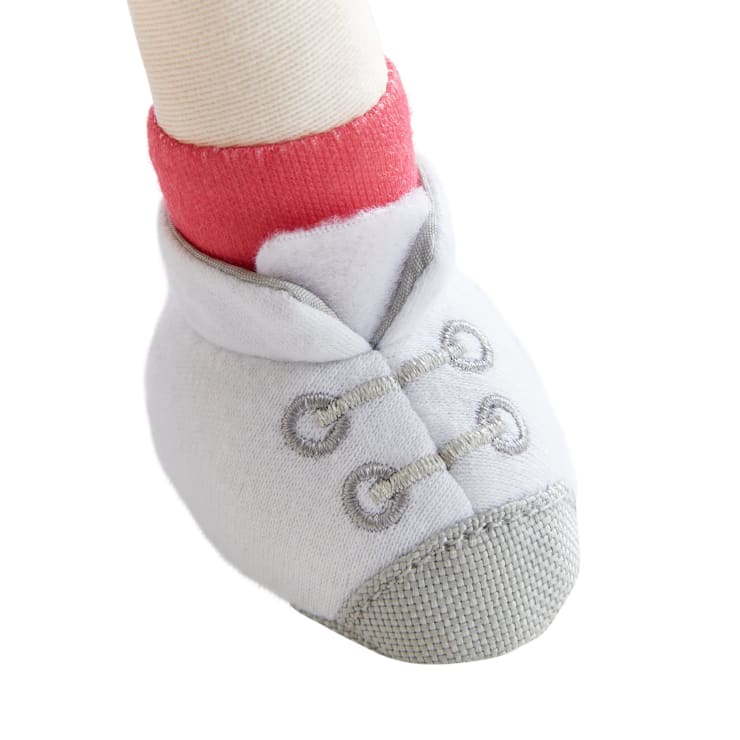 Nahaufnahme eines Babyfußes, der einen weißen Schuh aus weichem Stoff mit grauen Nähten und einer roten Socke darunter trägt. Der Schuh hat zwei dekorative Schlaufen und ist so gestaltet, dass er wie ein Turnschuh aussieht.