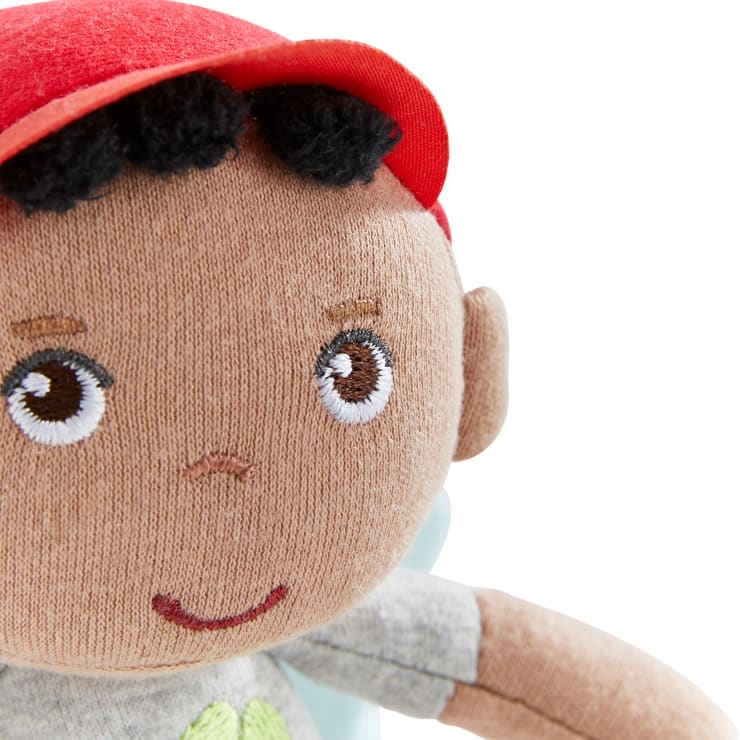Nahaufnahme einer Stoffpuppe mit brauner Haut, gestickten braunen Augen, einer kleinen Nase und einem kleinen Mund, schwarzem Garnhaar und einer roten Mütze. Die Puppe trägt ein graues Hemd mit einem grünen Muster. Der Hintergrund ist weiß.