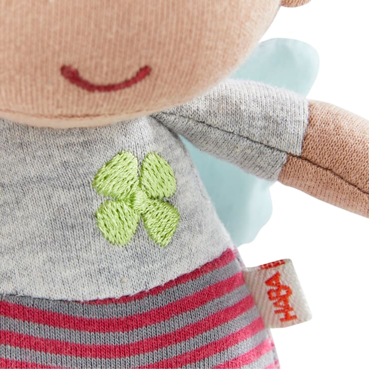 Eine Nahaufnahme einer Stoffpuppe zeigt einen Teil ihres Gesichts, ein graues Oberteil mit einem aufgestickten grünen Kleeblatt, eine rosa und grau gestreifte Hose und ein weißes Schild mit roter Aufschrift "HABA" an der Seite.