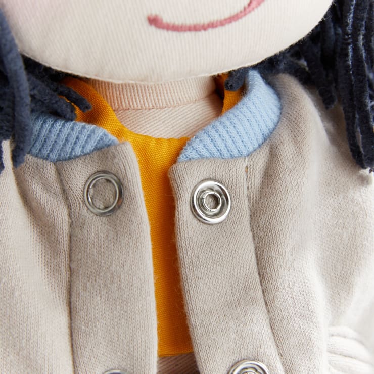 Nahaufnahme einer Stoffpuppe, die eine helle Jacke mit Metalldruckknöpfen, einen blauen Kragen und darunter ein gelbes Hemd trägt. Oben sind ein Teil des Gesichts der Puppe und dunkles Garnhaar zu sehen.