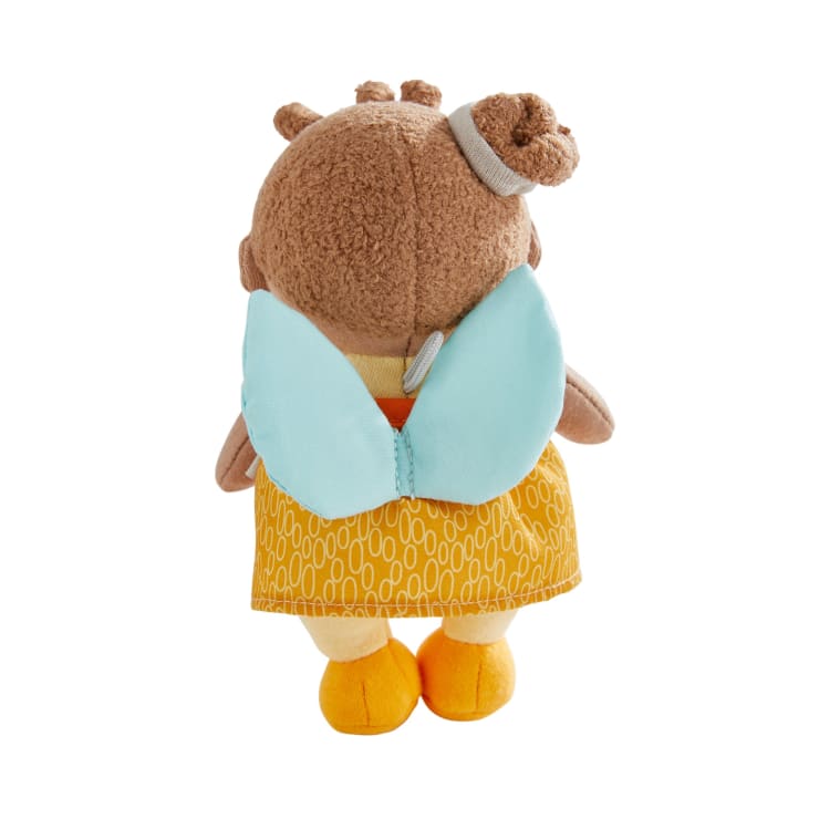 Eine von hinten gezeigte Plüschpuppe mit braunem Stoffhaar in einem Dutt, blauen Flügeln, einem gelben Kleid mit einem weißen ovalen Muster und gelben Schuhen. Der Hintergrund ist einfarbig weiß.
