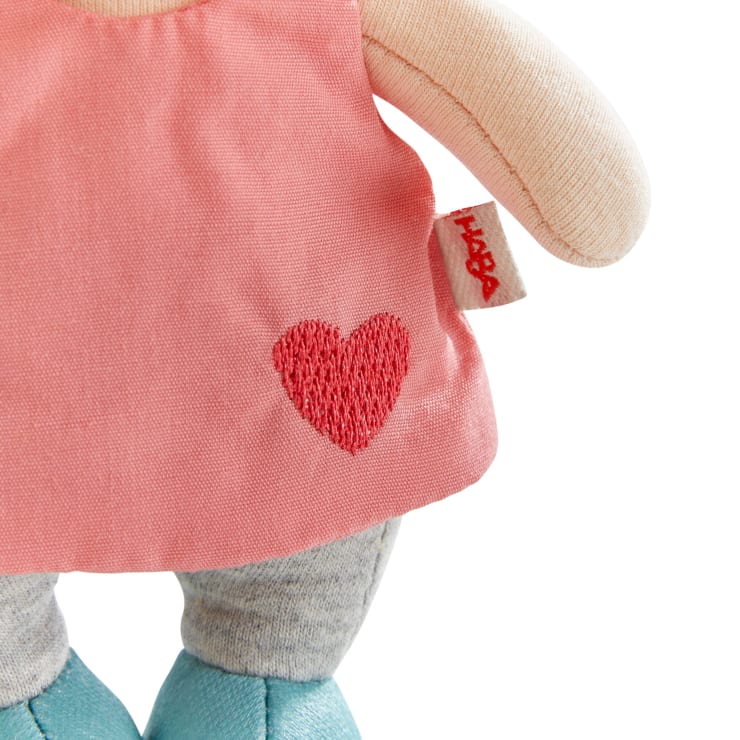 Eine Stoffpuppe, die ein rosa Kleid mit einem roten gestickten Herz, graue Leggings und blaue Schuhe trägt. Nur der Torso und die Oberschenkel der Puppe sind sichtbar.