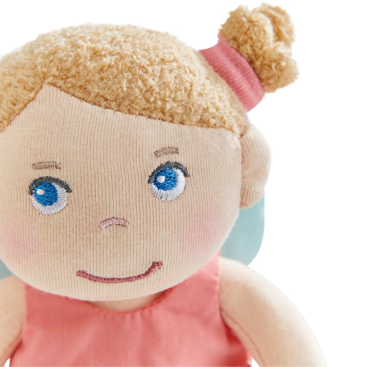 Eine Stoffpuppe mit blau gestickten Augen, hellbeiger Haut und blondem Garnhaar, das mit einem rosa Band zu einem Dutt gebunden ist. Die Puppe trägt ein rosafarbenes Outfit und hat ein leichtes Lächeln.