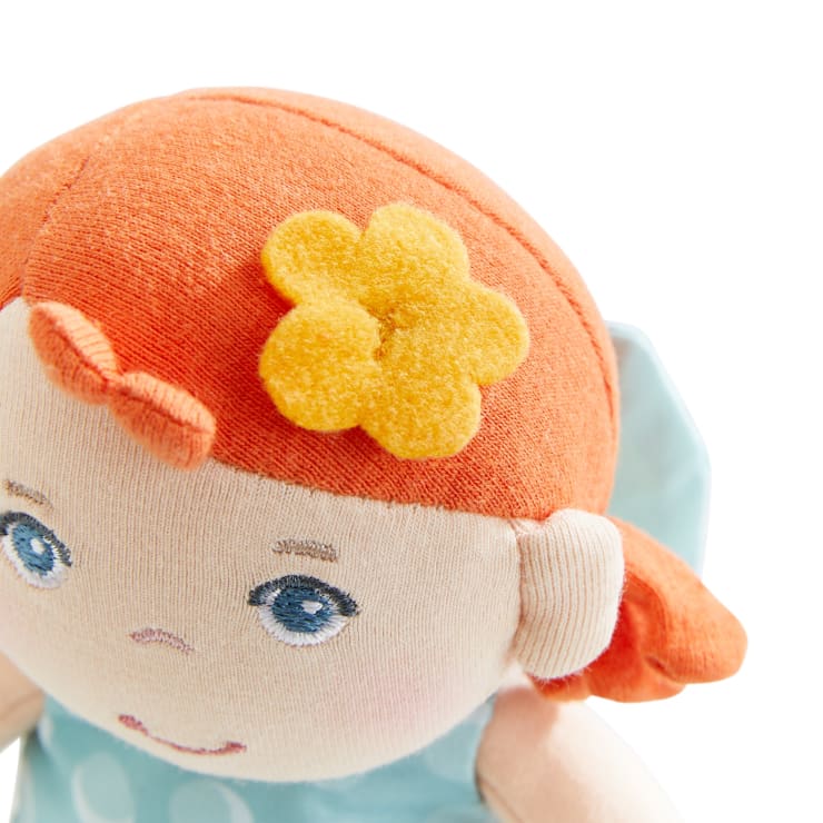 Eine Nahaufnahme einer Stoffpuppe mit orangefarbenen Haaren, blauen Augen und einem kleinen Lächeln. Die Puppe hat eine gelbe Filzblume im Haar und trägt ein hellblaues Kostüm. Der Hintergrund ist weiß.