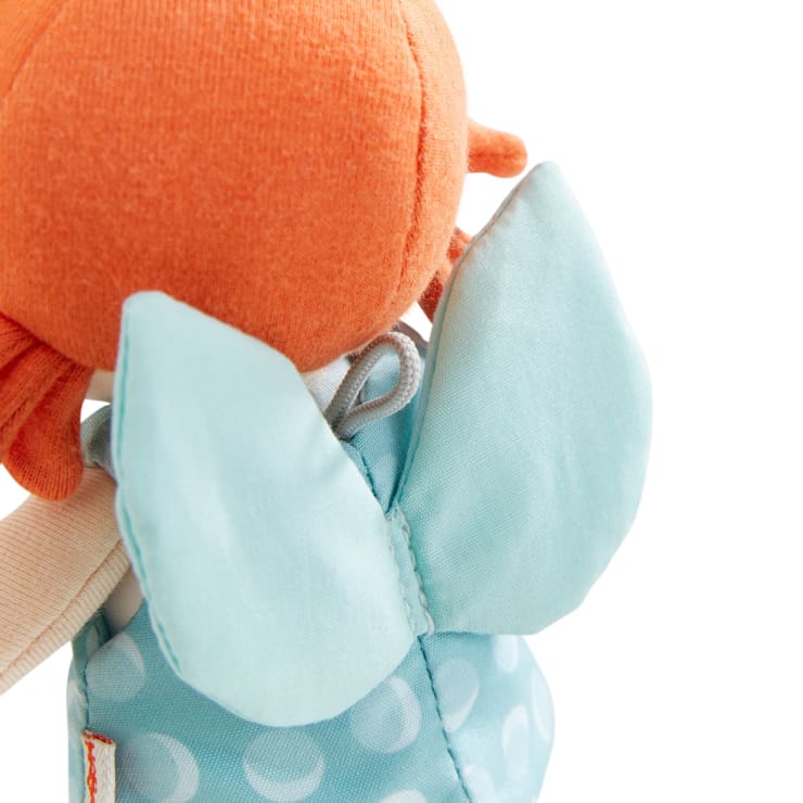 Eine Nahaufnahme einer Stoffpuppe mit orangefarbenen Haaren und hellblauen Flügeln. Die Puppe trägt ein blaues Kleid mit weißen Tupfen, und der Hintergrund ist weiß.