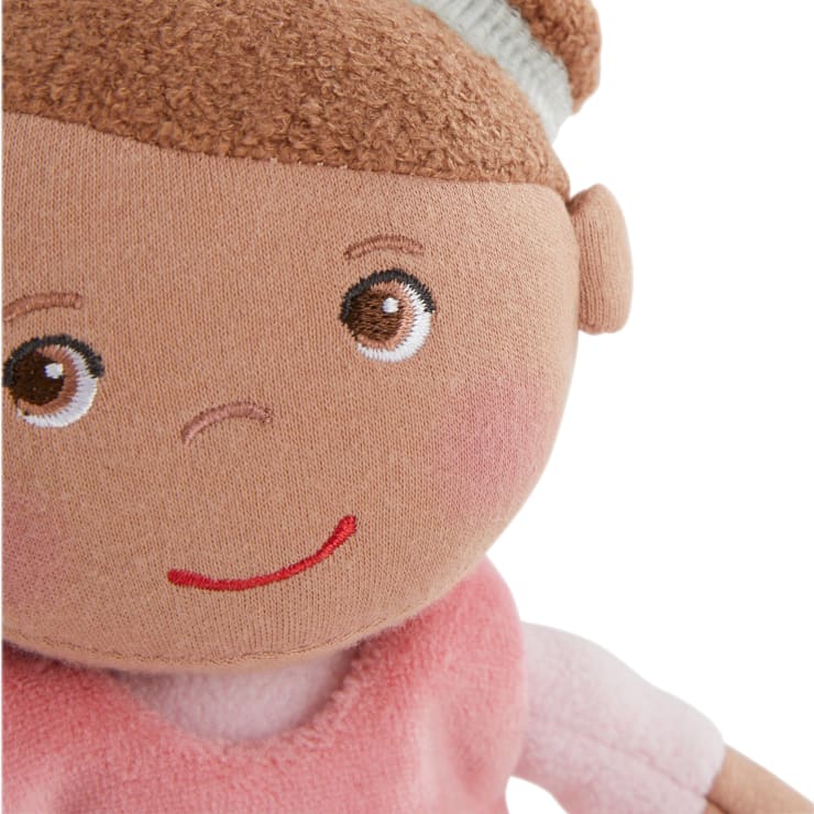 Nahaufnahme einer weichen Stoffpuppe mit braunen gestickten Augen, einem aufgestickten roten Lächeln, braunen Haaren und einem rosa Outfit. Der Kopf der Puppe ist leicht zur Seite geneigt.