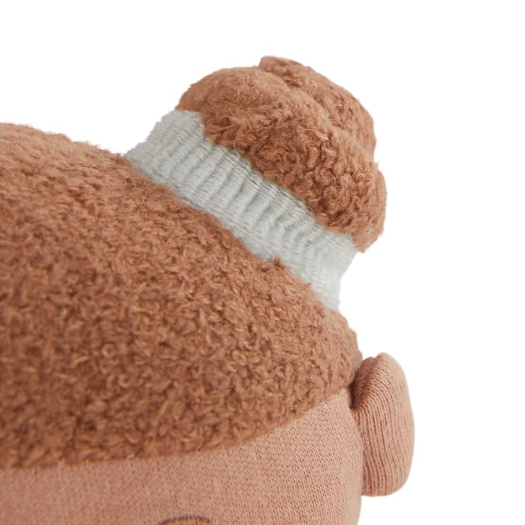 Nahaufnahme eines braunen Plüschpuppenkopfes mit strukturiertem braunem Haar, das oben zu einem Dutt frisiert und mit einem hellgrauen Band umwickelt ist, und einem sichtbaren Ohr auf der rechten Seite.