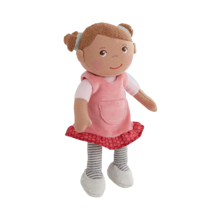 Eine weiche Stoffpuppe mit hellbraunem Haar in Zöpfen. Sie trägt ein rosa Kleid mit rotem Tupfensaum, grau-weiß gestreifte Leggings und graue Schuhe. Die Puppe hat gestickte Gesichtszüge und ein kleines Lächeln.