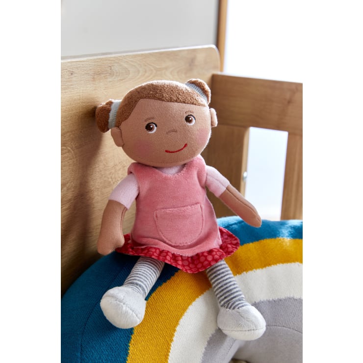 Eine Stoffpuppe mit braunen Haaren in Zöpfen, die ein rosa Kleid trägt, sitzt auf einem Bett neben einem blauen, gelben und weißen Kissen. Im Hintergrund sind ein hölzerner Bettrahmen und ein Fenster zu sehen.