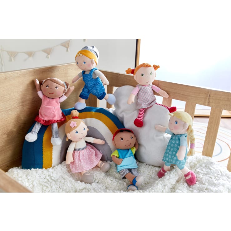 Sechs Stoffpuppen mit unterschiedlichen Haarfarben und Outfits sitzen und lehnen an einem Kissen und einem Regenbogenkissen in einem Holzbett, das mit einer weißen, strukturierten Decke gefüllt ist.