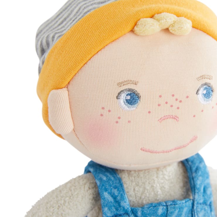 Eine Nahaufnahme einer weichen Stoffpuppe mit blauen Augen, rosa Sommersprossen und einem kleinen Lächeln. Die Puppe trägt einen gelb-grau gestreiften Hut und eine blaue Latzhose über einem cremefarbenen Hemd.