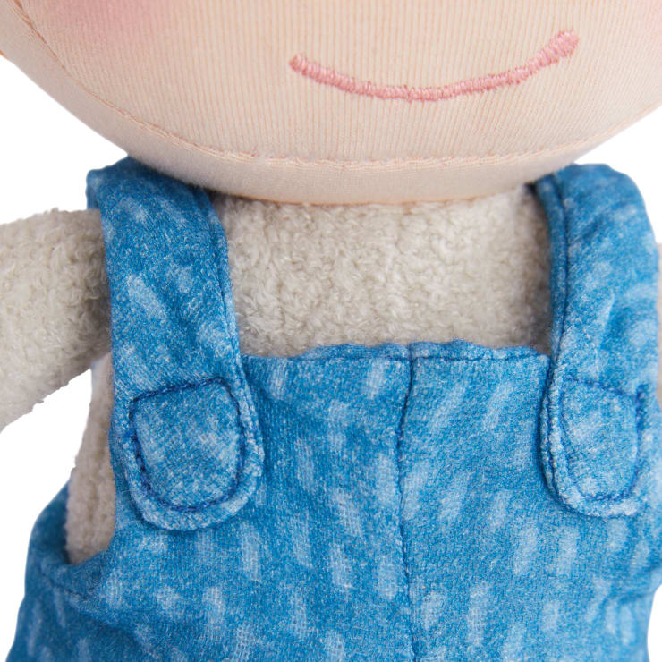Nahaufnahme einer Plüschpuppe, die eine blaue Latzhose in Jeansoptik und ein cremefarbenes strukturiertes Hemd trägt, das einen Teil des Puppengesichts mit einem einfachen aufgestickten rosa Lächeln zeigt.
