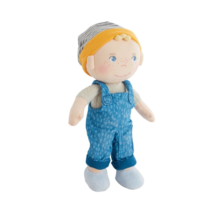 Eine Stoffpuppe mit heller Haut, blauen Augen und Sommersprossen trägt ein gelbes Stirnband, einen grau gestreiften Hut, eine blaue Latzhose, ein beiges Hemd und hellblaue Schuhe und steht vor einem weißen Hintergrund.