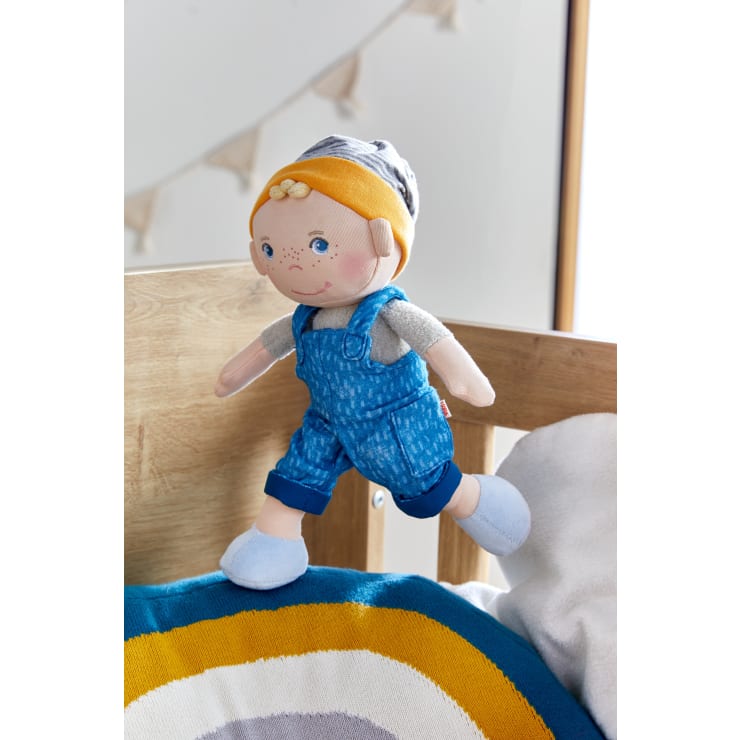 Eine Stoffpuppe mit gelbem und grauem Hut, grauem Hemd, blauer Latzhose und blauen Schuhen sitzt auf der hölzernen Kante eines Kinderbetts mit einem bunten Kissen und einer unscharfen Wimpelkette im Hintergrund.